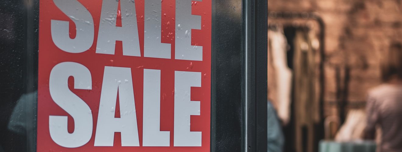 Red sale sign on a glass door with interior view of a clothing store.