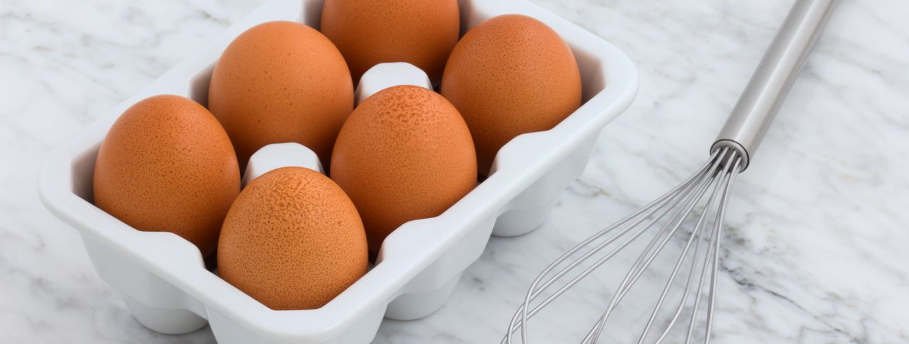 Six brown eggs in a carton and a metal whisk on a marble countertop. Perfect for cooking concepts.