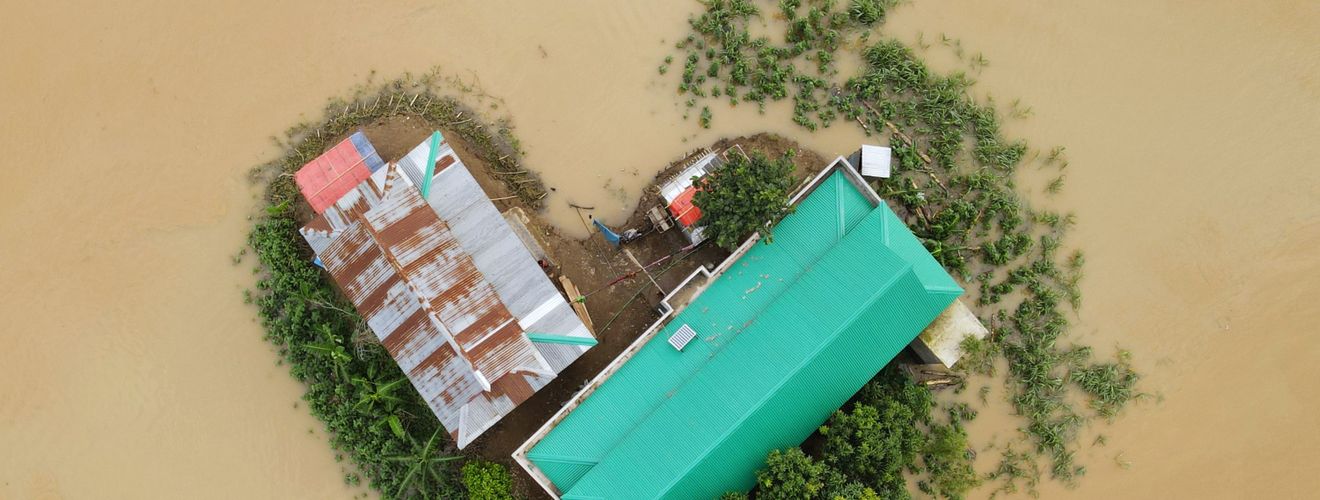 Top-down view of a flood-affected area in Bangladesh showing buildings and surrounding submerged land.
